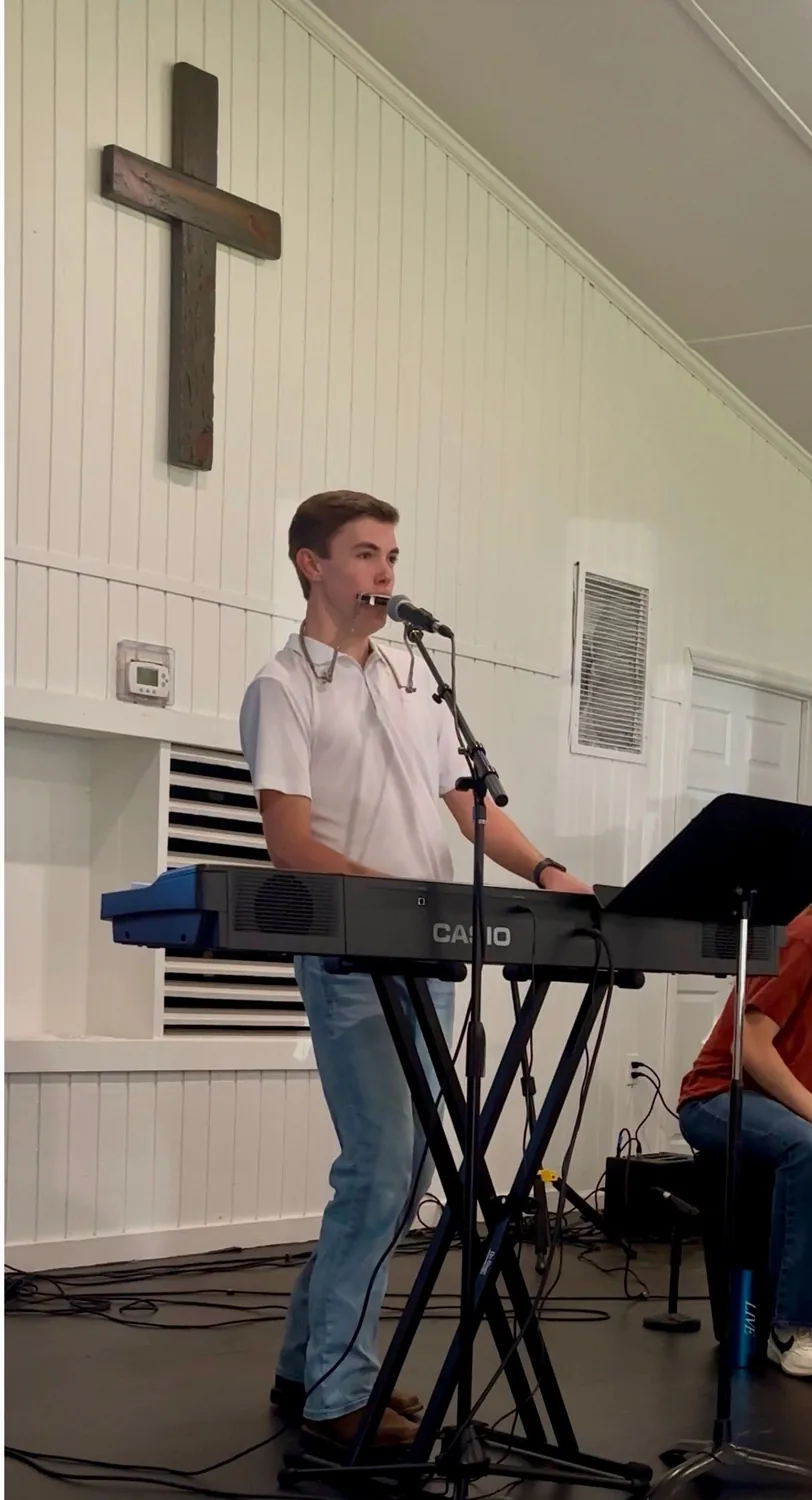 AJ Robinson playing keyboard and harmonica during worship, a wooden cross behind him